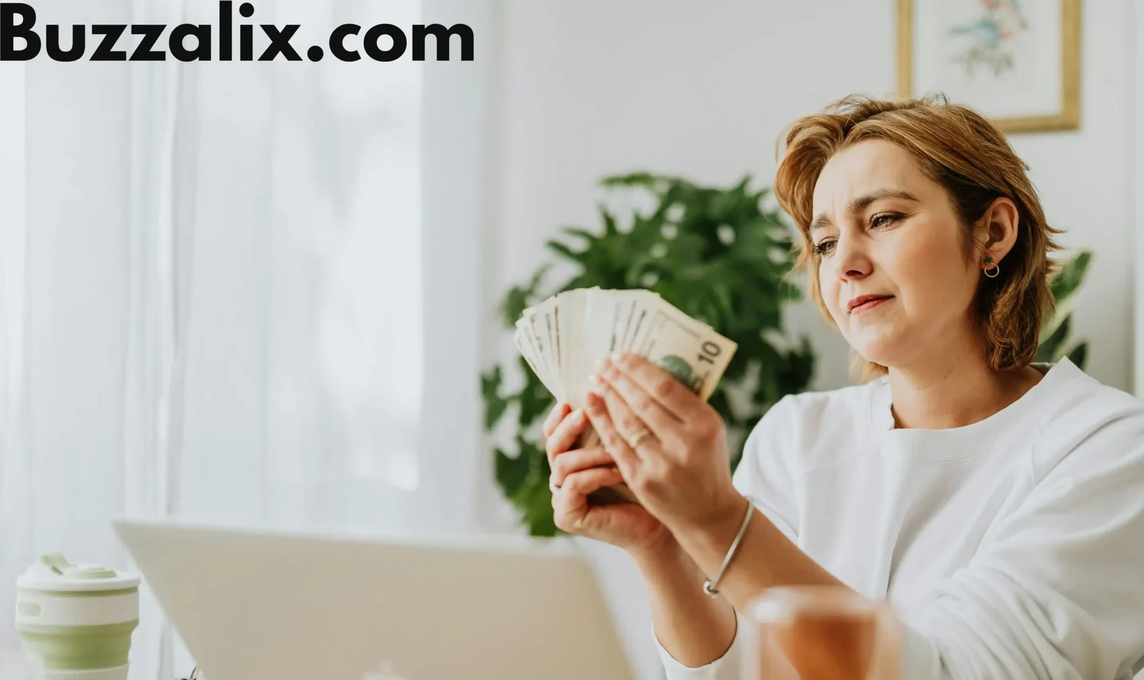 A woman sitting at her desk with a laptop, counting money while researching tech salaries, representing income growth in the 2025 Tech Salary Guide by Buzzalix.